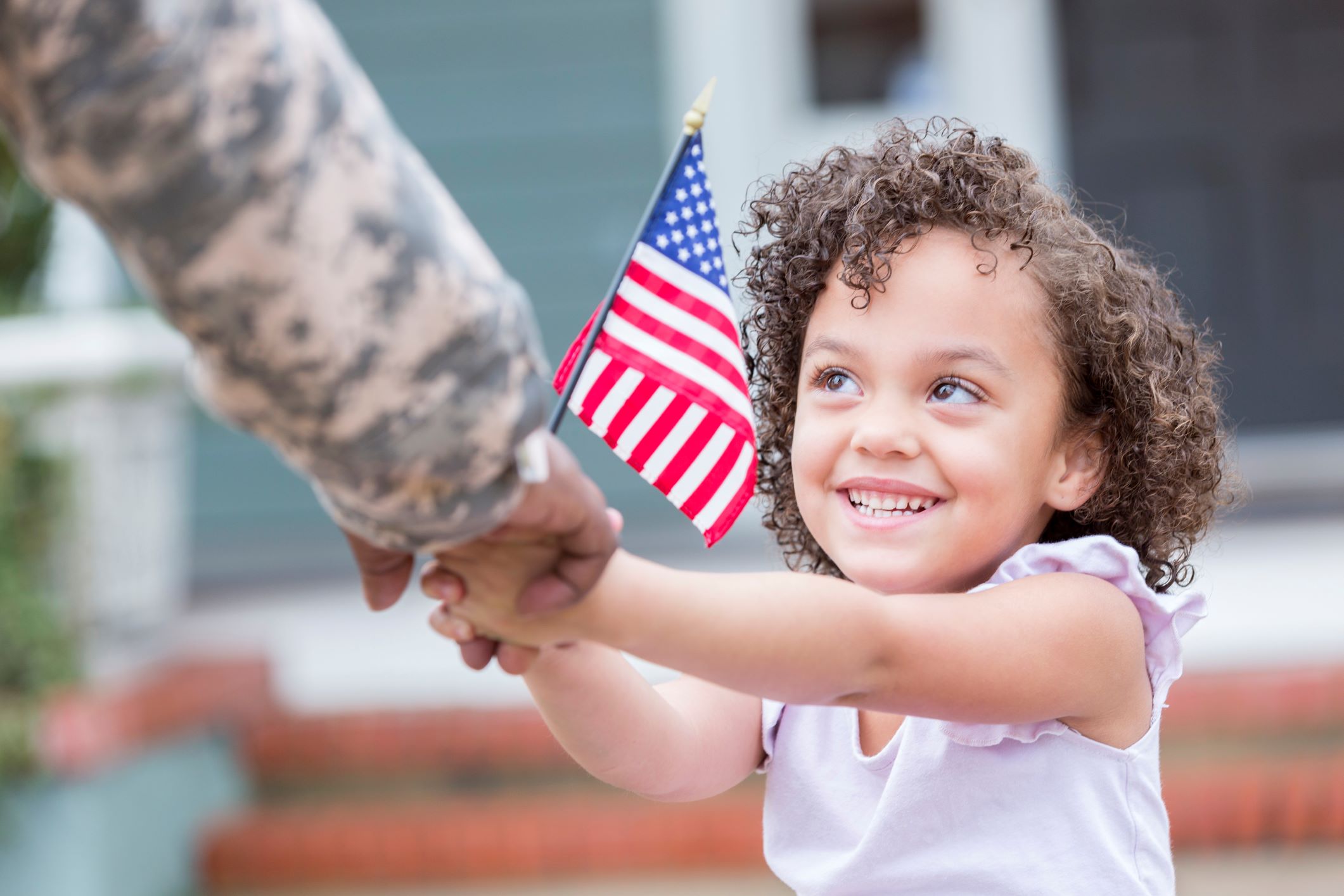 a young girl holds the hand of her father who is in the Army. he's wearing his army uniform and they are holding a mini american flag together VA Mortgage Loans US Army Garrison