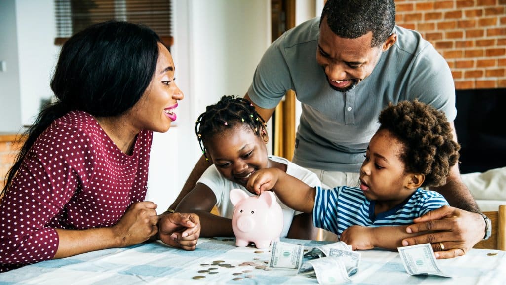 Parents teaching their two young children the importance of saving and having the children put money into a piggy bank.