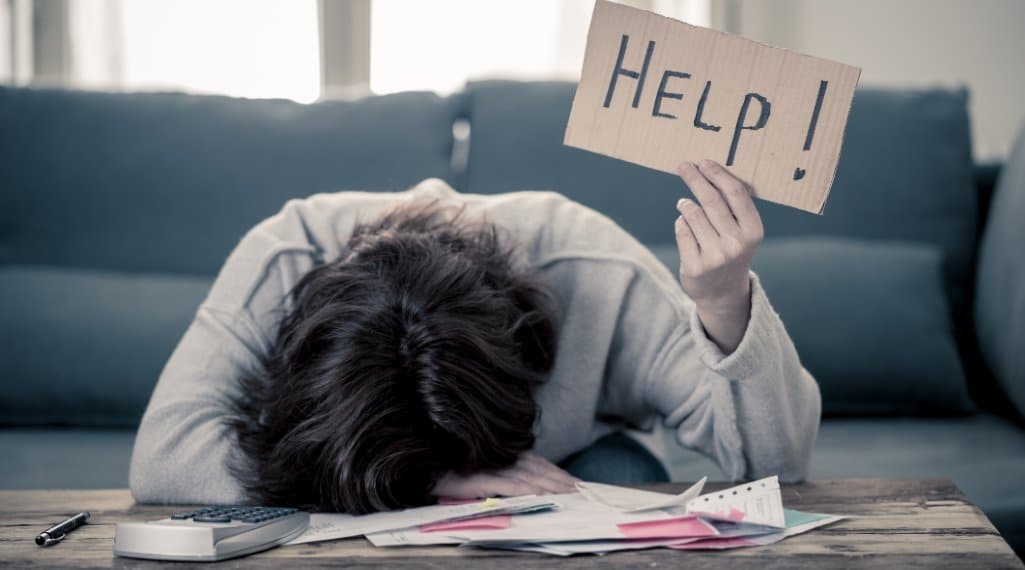 Woman with her head face down on a table full of tax forms holding a cardboard sign with the word "HELP" written on it.