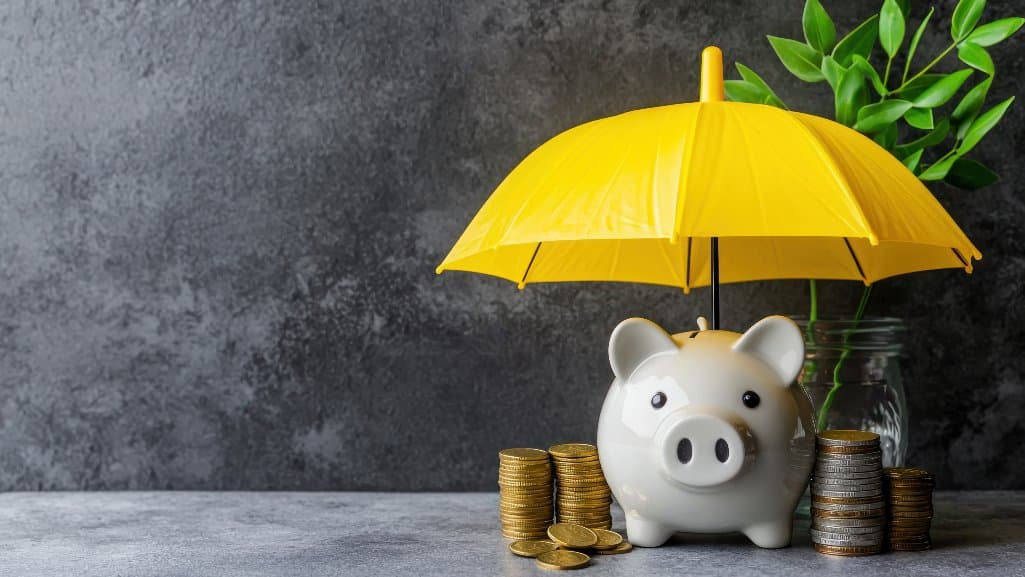 Stacks of coins next to a piggy bank all under the protection of a yellow umbrella.
