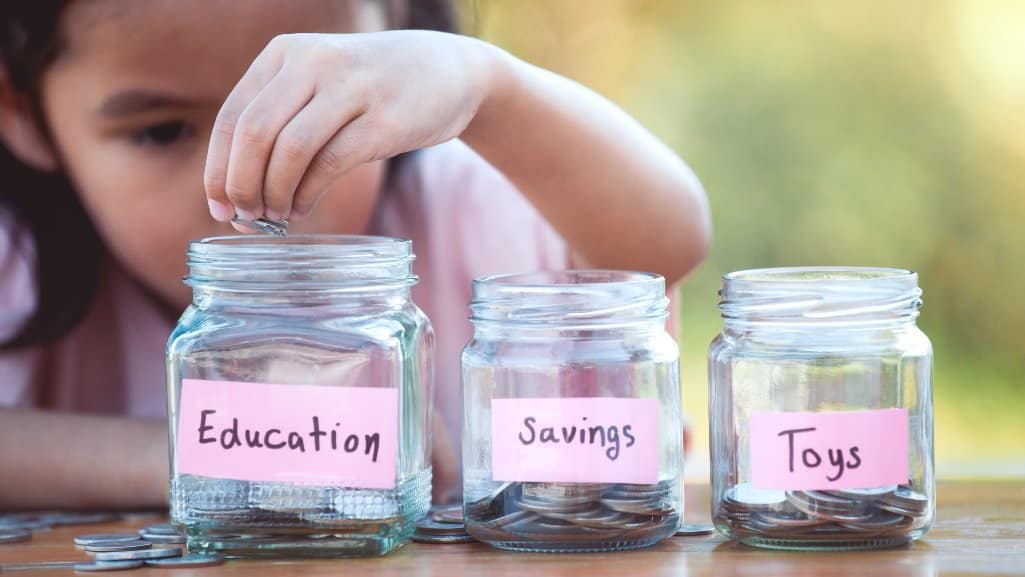 Child putting coins in glass jars labeled "Education," "Savings," and "Toys."