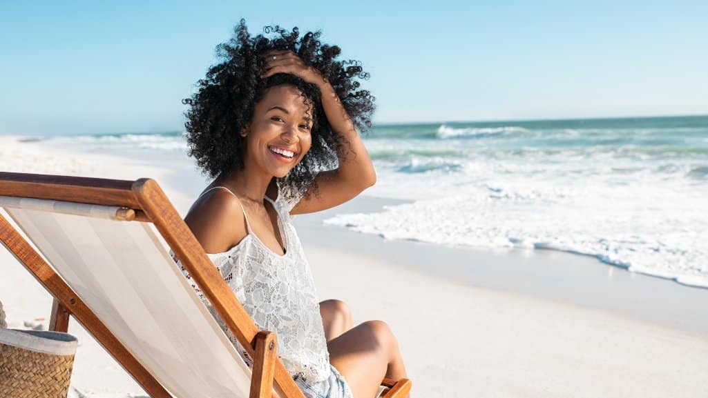 Woman relaxing on the beach in a lounge chair, turning to face the camera and smiling.
