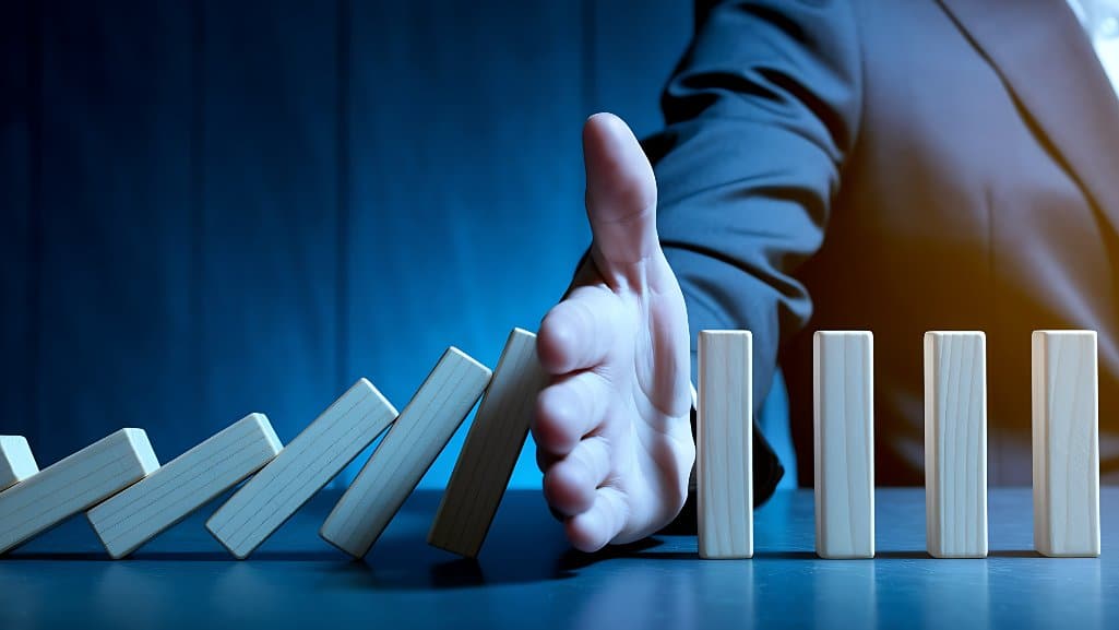 A businessperson’s hand stops a row of falling dominoes, symbolizing proactive fraud intervention and risk prevention.