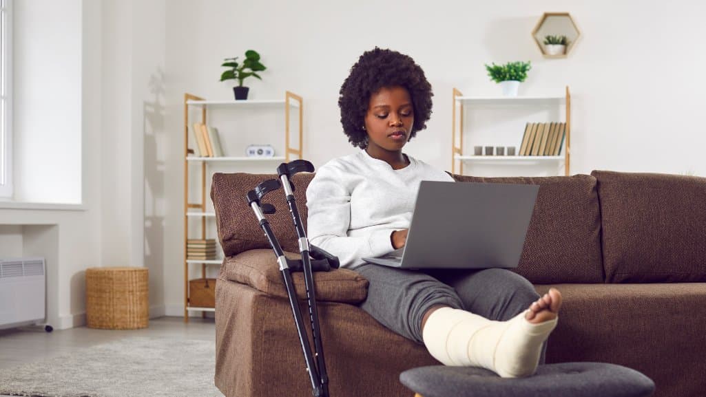 Woman with a cast on her leg, sitting on her couch, resting her broken leg on the ottoman, while shopping online for a debt consolidation loan.