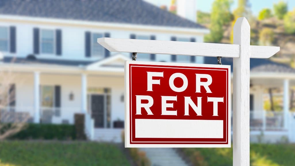Red and white For Rent sign in front of a suburban house, symbolizing online rental listings and scams.