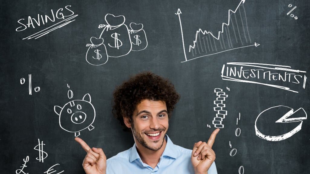 Smiling man pointing upward in front of a chalkboard covered with financial doodles, including savings bags, a piggy bank, charts, and investment symbols — representing learning about money and financial literacy.