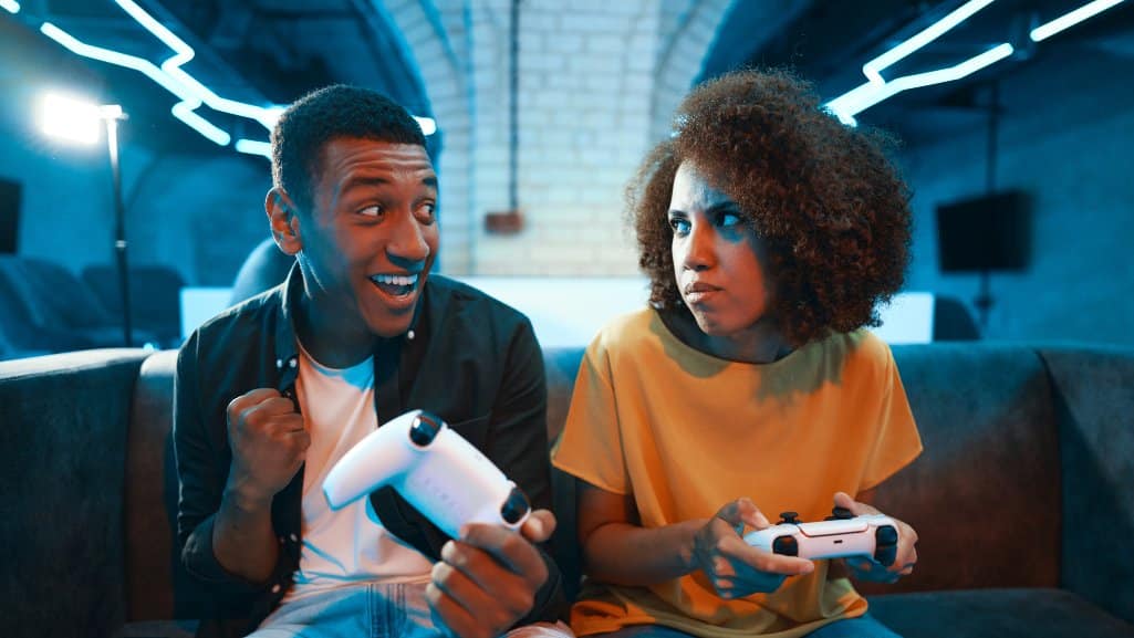 Two gamers sitting on a couch with wireless controllers, one celebrating a win while the other looks frustrated, highlighting how video games can have real-world impacts.
