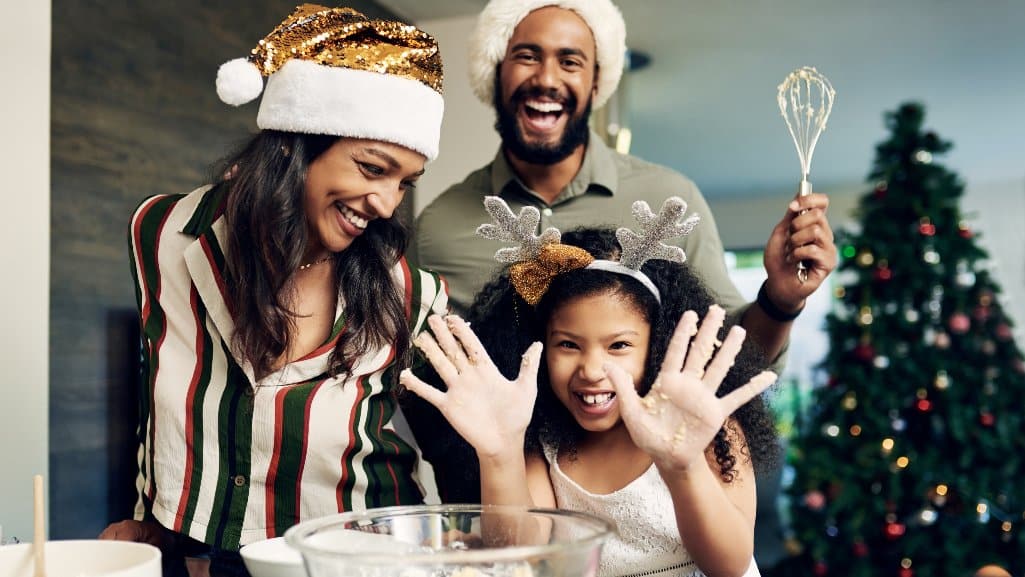 Family baking holiday treats together in the kitchen, wearing festive hats, with a Christmas tree in the background.