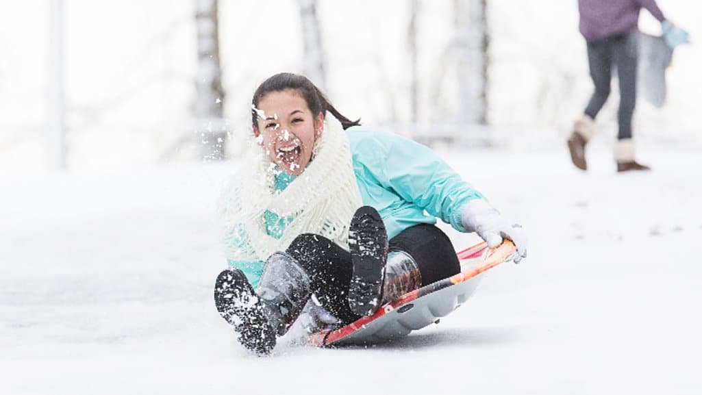 Smiling teen sledding down a snowy hill, kicking up snow while enjoying a winter day.