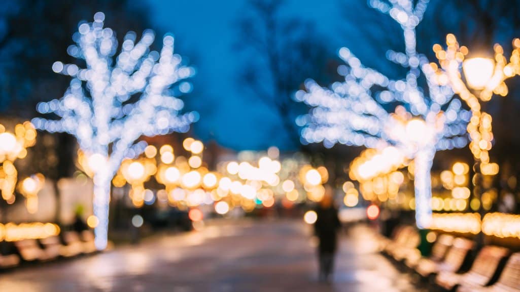 Blurry view of a city walkway at night with trees and lampposts covered in holiday lights.