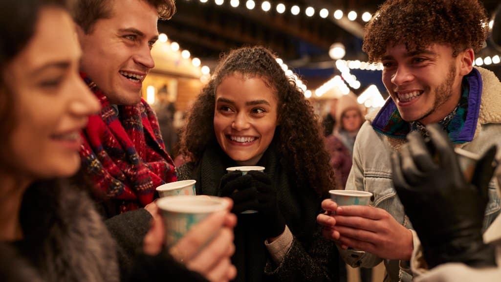 Group of friends at an outdoor holiday market, smiling and holding hot drinks under string lights.