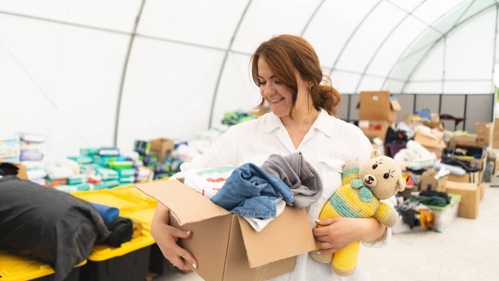 Volunteer carrying a box of donated clothes and a stuffed toy inside a community donation center.