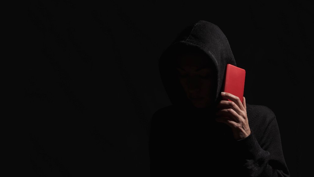 A shadowy person in a hooded sweatshirt holding a red smartphone against a dark background, representing an account takeover scam.