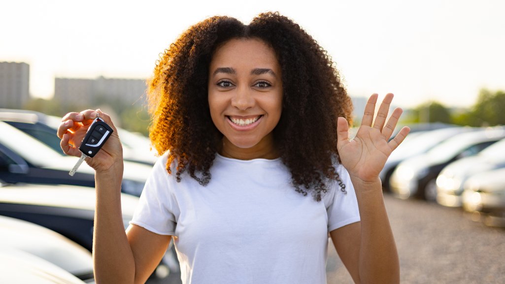 Smiling young woman in a used-car sales lot holding up the key to her new car she purchased with an auto loan from People Driven Credit Union.