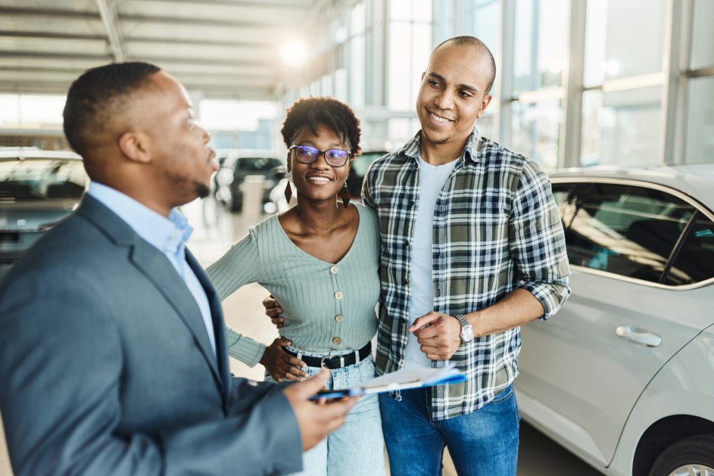 Happy couple talking about vehicle loan plans with a salesman in car showroom.